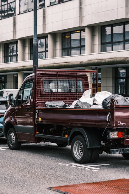 A burgundy flatbed pickup truck parked on a city street in front of a modern office building with large glass windows and concrete exterior panels. The truck's cargo bed is filled with various bags and bundles of mixed waste materials, including black and white plastic trash bags, some of which appear partially open, revealing indistinct contents. A metal safety grille is mounted on the cab’s rear window, providing protection during transport. The surrounding environment includes parked vehicles along the curb and a tall street lamp nearby. The scene reflects an everyday urban setting with informal waste collection possibly managed through private waste removal services, such as those offered by House Clearance Uxbridge, that handle on-site rubbish removal and bulky waste disposal.