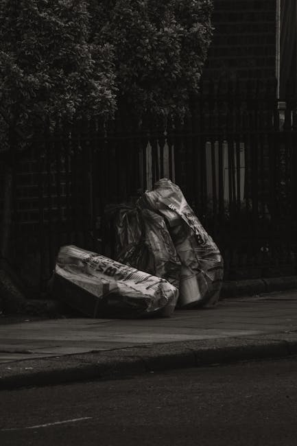 Two large black plastic bags filled with household or garden waste are placed on the pavement near a dark metal fence, which appears to be part of a residential property. The bags are slightly crumpled and reflective, with some text and logos visible on their surfaces. Behind the fence, dense foliage of trees or bushes is visible, creating a natural backdrop. The scene is illuminated by soft, natural lighting, suggesting an outdoor setting during daytime. The bags are positioned close to the curb, indicating they are awaiting collection, potentially by private rubbish removal services such as those offered by House Clearance Uxbridge. The environment appears tidy aside from the waste bags, emphasizing the need for organized rubbish removal or waste management in the local area, supporting the concept of alternative waste handling outside of municipal collection processes.