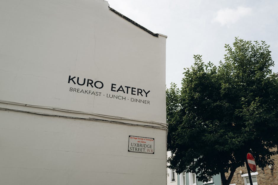 A white exterior wall of a building displaying a sign with the text 'KURO EATERY' and smaller wording beneath indicating 'BREAKFAST - LUNCH - DINNER'. To the right of the signage, a street name sign reads 'UXBRIDGE STREET W8' and is mounted on the wall, slightly below the larger sign. Adjacent to the building, a leafy green tree extends its branches outward, partially obscuring the corner of the building. The sky above is overcast with a light grey tone, providing neutral natural lighting. In the lower right corner, a traffic sign is visible, indicating a no-entry or restriction zone, and is partially obscured by the tree. The scene exudes a quiet, urban atmosphere typical of a residential or commercial district, with the signage suggesting the presence of a cafe or restaurant. The image's context relates to local street surroundings, which might associate with nearby waste disposal or collection points, consistent with the themes of rubbish removal and private waste handling often encountered in urban settings. As part of a UK-based waste management services page, the image subtly reflects the typical environment where such services may be required, though no waste or removal activities are visually depicted in this scene.