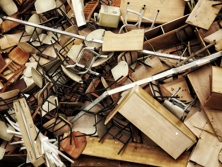 A chaotic pile of discarded office furniture and equipment, including numerous wooden desks, metal and wooden chairs with various finishes, some with black or wooden frames and upholstery, all stacked and scattered across a flat surface. The desks are of different sizes and styles, with some having attached storage drawers and others plain surfaces, with finishes ranging from light to dark wood tones. Chairs are intertwined, with some upside-down, leaning, or toppled, showing plastic, metal, and wood components. Visible in the background are additional furniture items, including a small wooden cabinet and a white appliance, possibly a microwave or small refrigerator. There are metal rods or supports, possibly parts of shelving or framing, running diagonally across the scene. The environment appears to be indoors, in a storage or clearance area used by House Clearance Uxbridge for rubbish removal, illustrating a collection of items prepared for disposal or private waste handling, characteristic of an on-site clearance operation for bulky refuse dismissal.