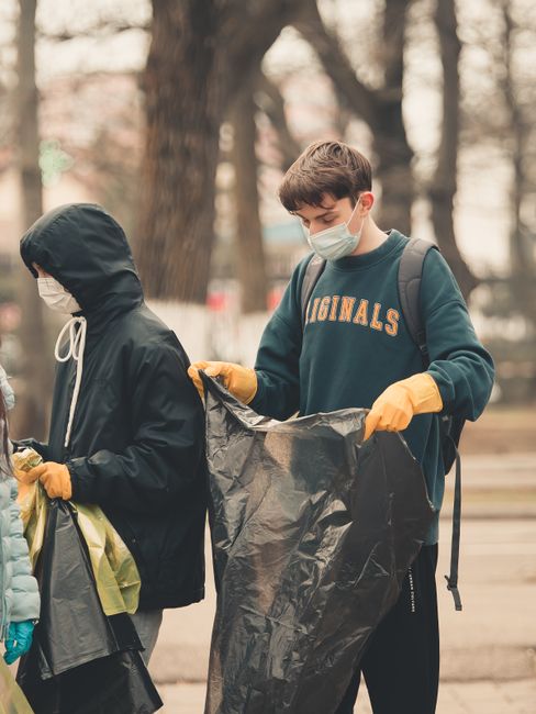 Two young individuals participating in a community rubbish clearance activity outdoors during daytime, both wearing protective face masks and yellow gloves. The young man on the right, with light brown hair, is dressed in a dark sweatshirt with the word 'ORIGINALS' printed across the chest, and is holding a large black plastic bag, appearing to be ready for waste collection. The person on the left, wearing a black hooded jacket with the hood up and a white face mask, is also holding a bag, seemingly engaged in rubbish sorting or collection. They are situated on a paved area with several mature trees in the background, suggesting a park or similar green space, with a natural, slightly blurred environment. This scene reflects an organised on-site rubbish removal effort, consistent with independent waste handling or community clean-up activities, which House Clearance Uxbridge often supports through professional waste removal services. The overall lighting is natural, with diffused daylight highlighting the textures of their clothing and the surrounding environment, emphasizing a focus on environmental responsibility and community engagement in waste management.