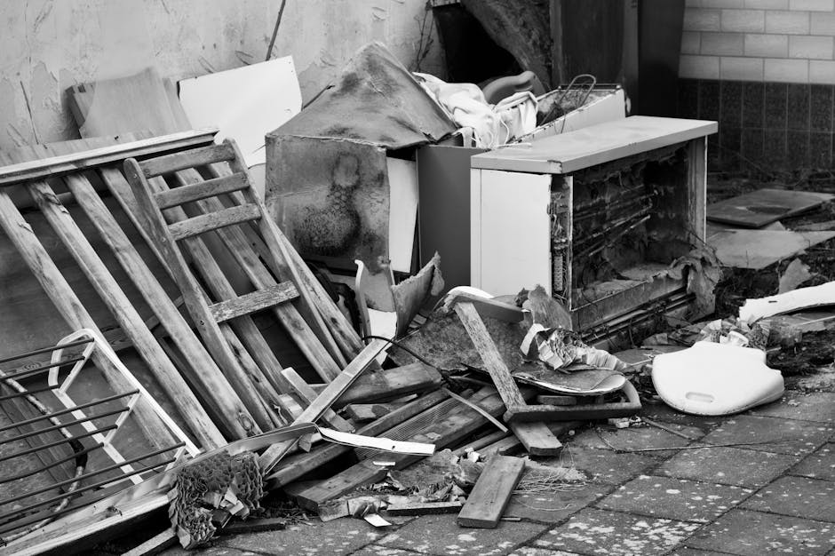 A chaotic pile of discarded office furniture and equipment, including numerous wooden desks, metal and wooden chairs with various finishes, some with black or wooden frames and upholstery, all stacked and scattered across a flat surface. The desks are of different sizes and styles, with some having attached storage drawers and others plain surfaces, with finishes ranging from light to dark wood tones. Chairs are intertwined, with some upside-down, leaning, or toppled, showing plastic, metal, and wood components. Visible in the background are additional furniture items, including a small wooden cabinet and a white appliance, possibly a microwave or small refrigerator. There are metal rods or supports, possibly parts of shelving or framing, running diagonally across the scene. The environment appears to be indoors, in a storage or clearance area used by House Clearance Uxbridge for rubbish removal, illustrating a collection of items prepared for disposal or private waste handling, characteristic of an on-site clearance operation for bulky refuse dismissal.